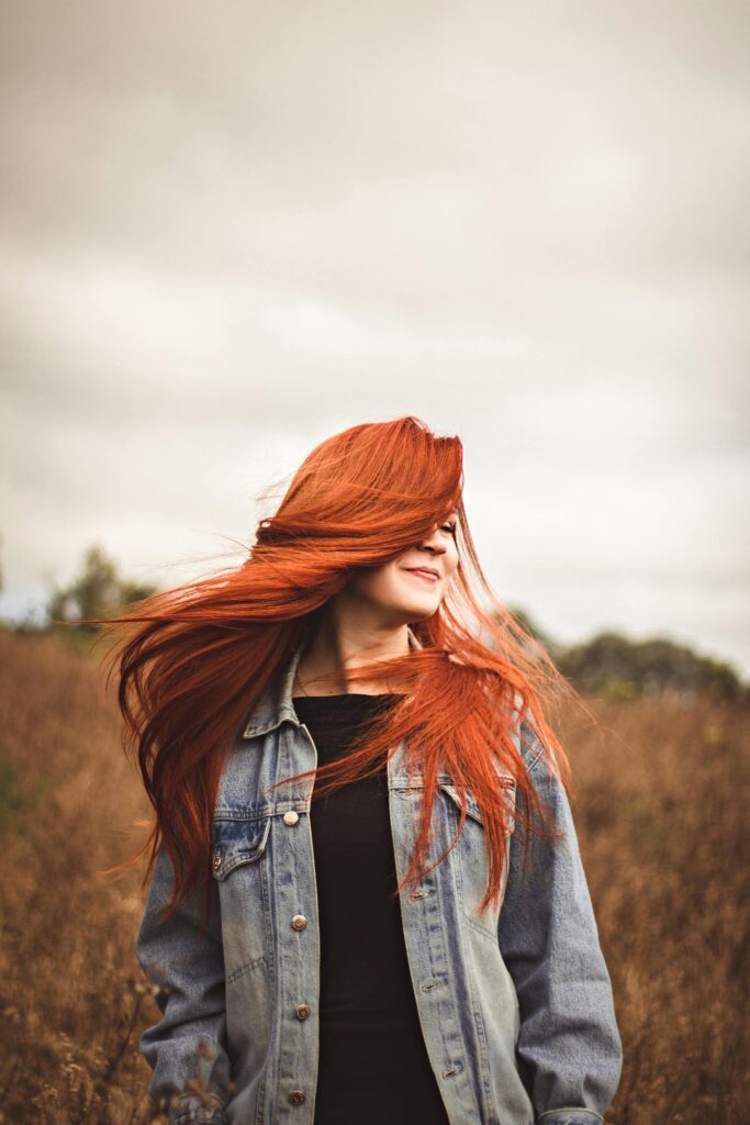 Smiling woman with red hair enjoys a breezy day in a scenic autumn field.