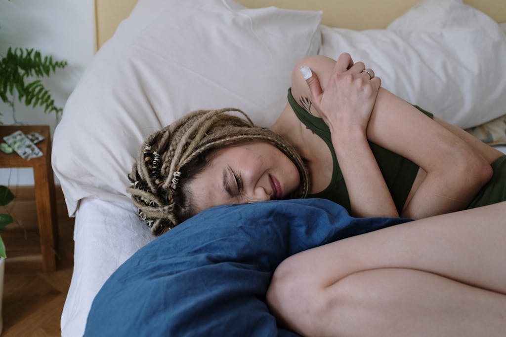 A woman with dreadlocks lying in bed holding her stomach in pain, possibly due to cramps.