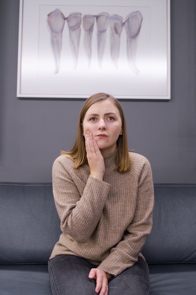 Woman in a brown sweater holding her cheek, indicating pain, in a dental clinic setting.