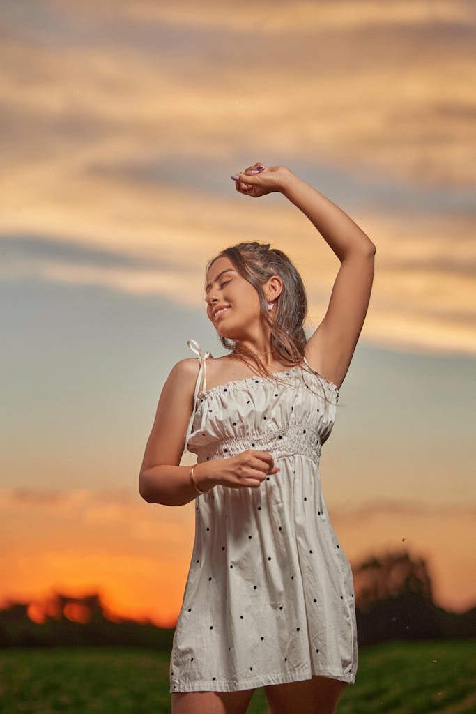 Young woman posing playfully in a white dress during a vibrant sunset.